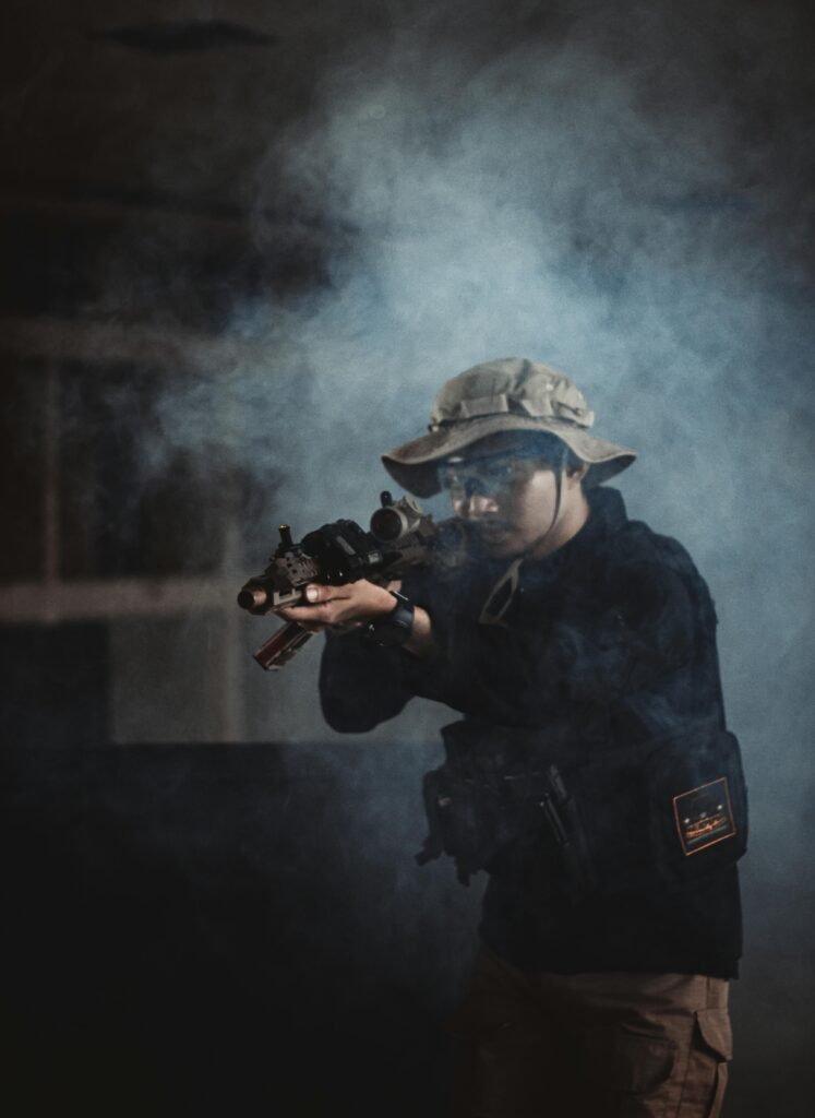A person in tactical gear with an airsoft rifle in a smoky indoor setting, playing a mock combat scenario.